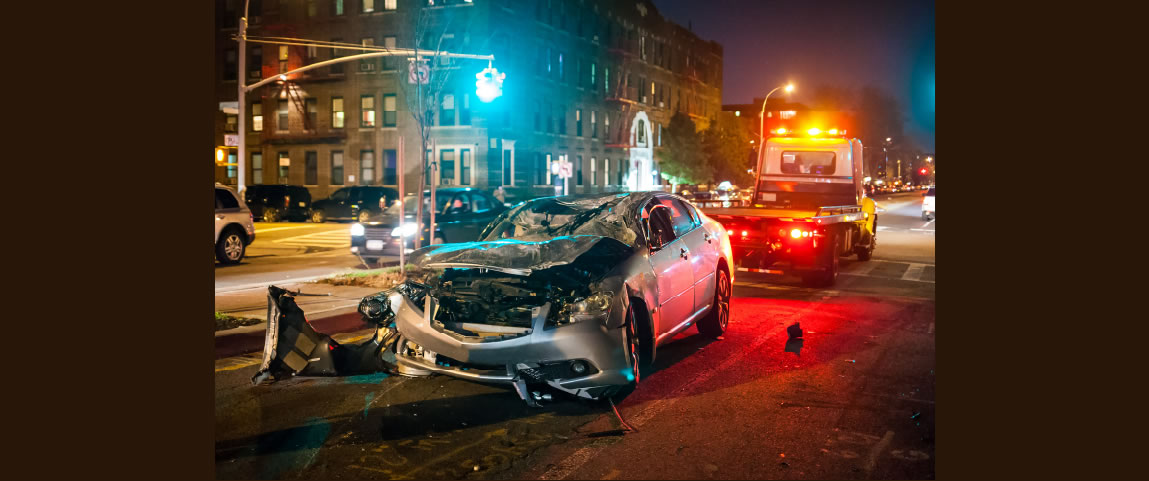 Severely damaged silver sedan after a car crash at nighttime, the front end of the vehicle is crushed and debris scattered on a city street. A tow truck with flashing yellow and red lights is parked behind the wreck while vehicles travel carefully next to the incident.